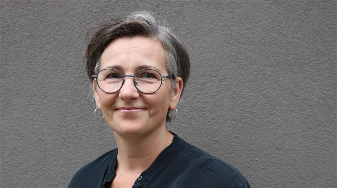 A woman with dark hair, glasses and a black blouse stands in front of a dark grey wall.
