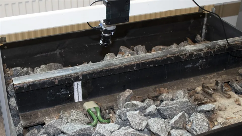 En bit räls i en pallkrage, omgiven av sten och ett clips är synligt. Ovanför syns en kamera som är riktad mot clipset.