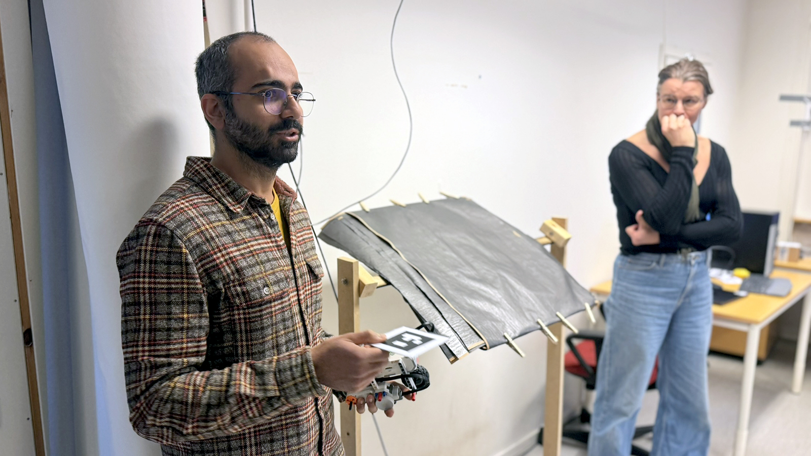 A man stand up and hold an object as he speaks. A woman standing in the background listening.