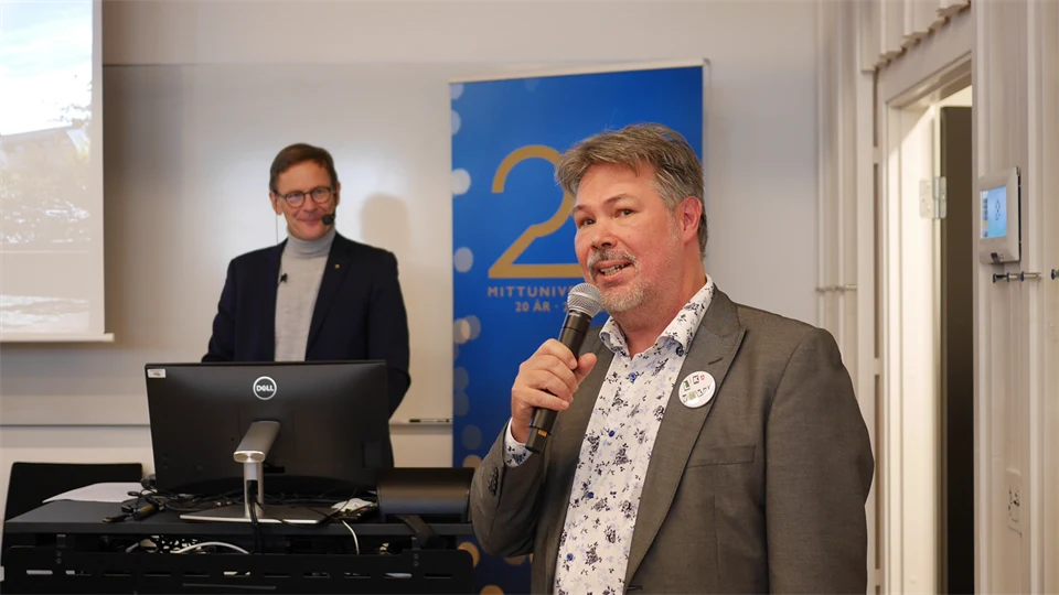 En man i grå kavaj och mönstrad skjorta står bredvid ett podium och håller i en mikrofon.