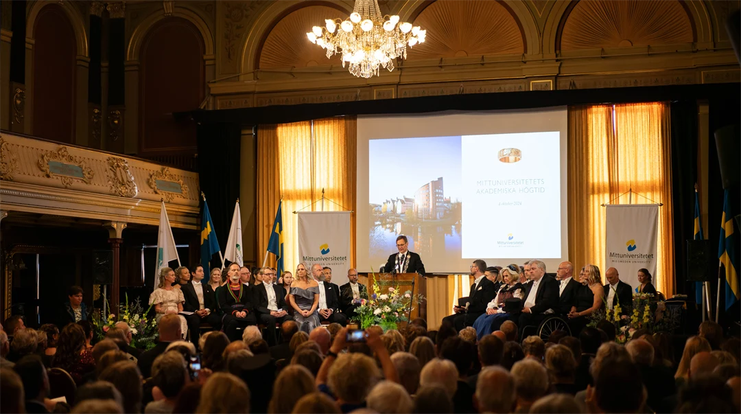 En finklädd man står i en talarstol och talar framför en publik.