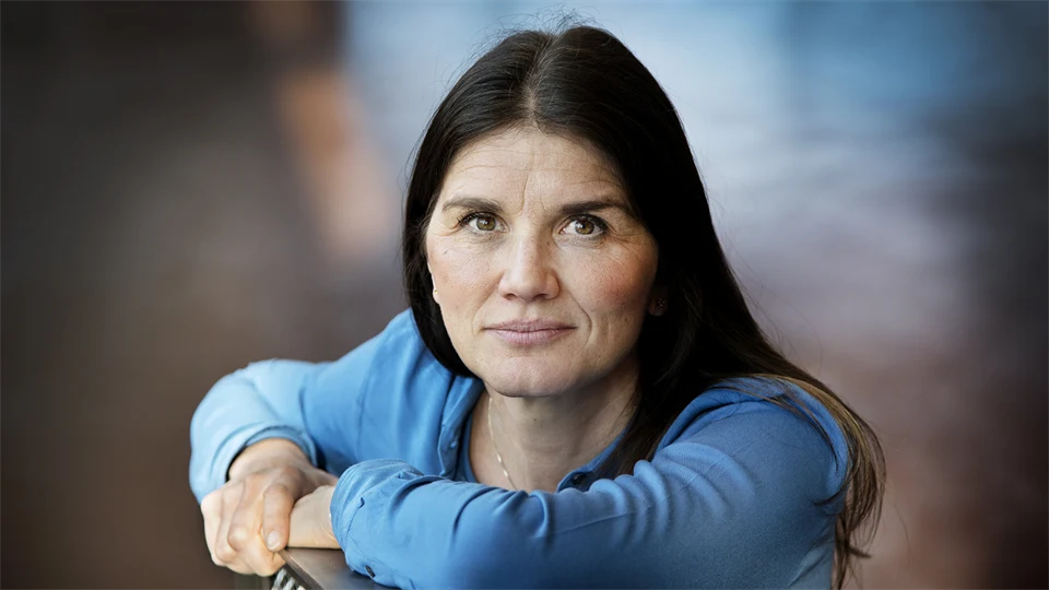 A woman with dark long hair, brown eyes and a blue sweater is leaning against the back of a chair and looking into the camera.
