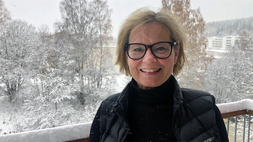 En kvinna med glasögon som ler. Träd med snö i bakgrunden