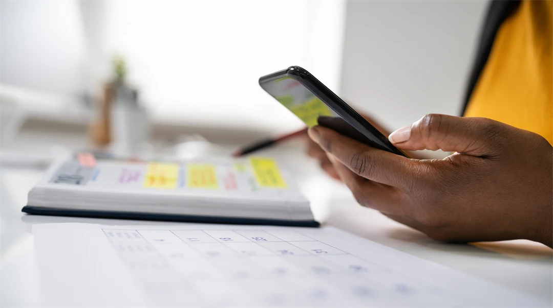 A hand is holding a mobile phone and a calendar is lying on a table.