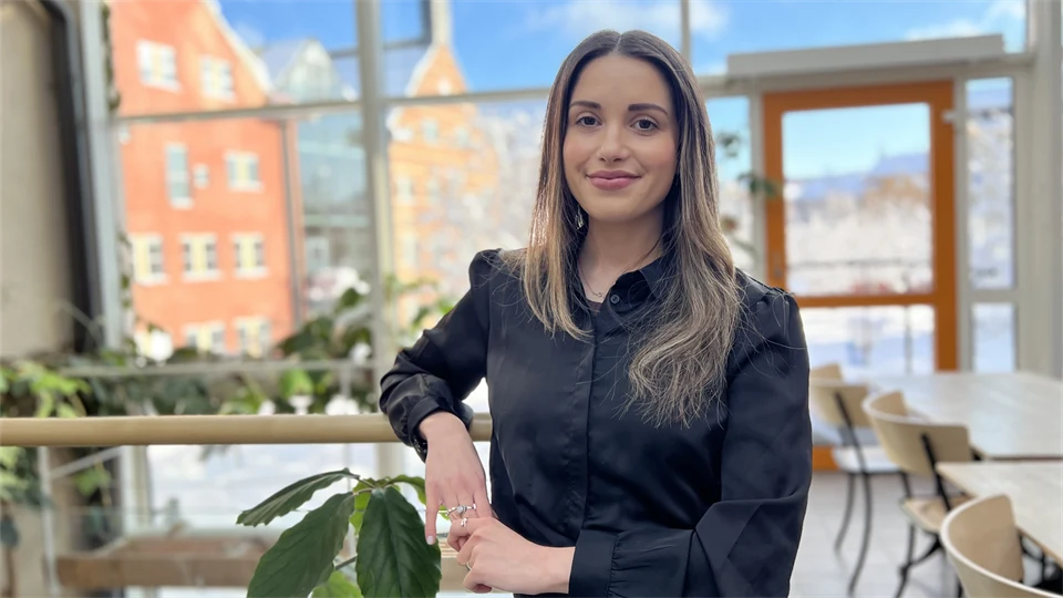 En kvinna tittar in i kameran och ler. Bakom hennes finns en glasvägg som visar husfasaderna utanför. 