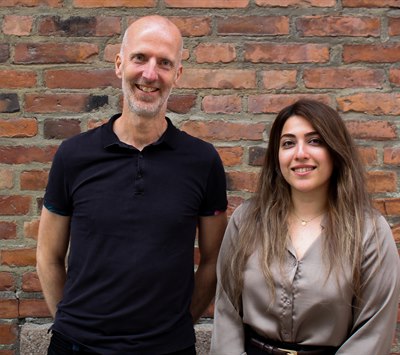 En man i svart pikétröja och en kvinna  i brun blus står bredvid varandra framför en tegelvägg. 