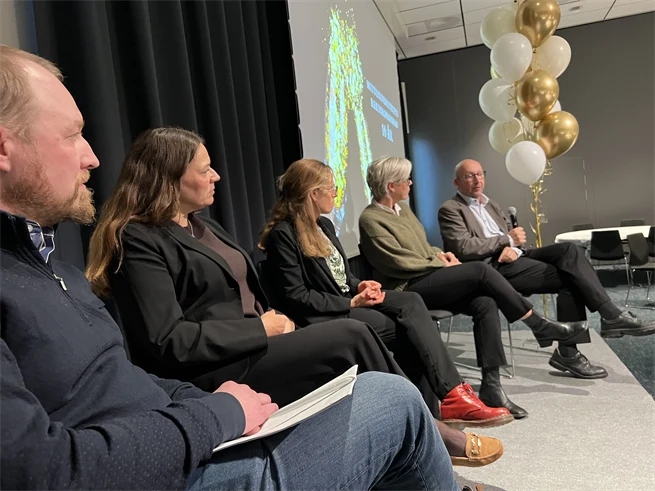 En scen med fem personer sittandes på stolar. I bakgrunden syns guldiga och vita ballonger.