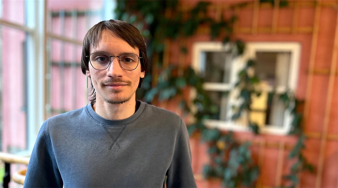 A man with brown hair, glasses and a grey jumper stands in front of a reddish-brown wall covered in climbing green plants. 