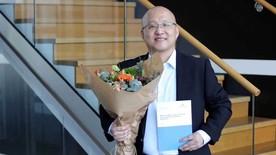Doctoral student Ye Xu stands by a staircase and holds a bouquet of flowers and his dissertation.
