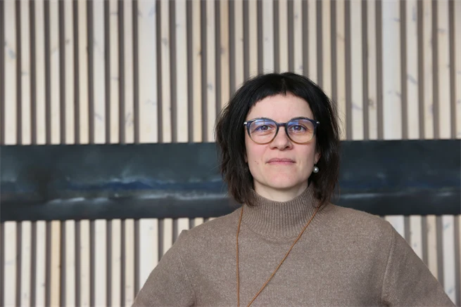 Allvarlig kvinna med glasögon framför träpanel.