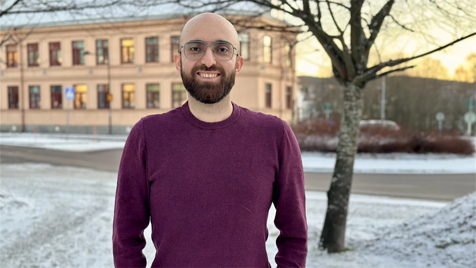 Smiling man with a shaved head, brown beard, and glasses. He is wearing a burgundy sweater and standing outdoors. The season is winter.