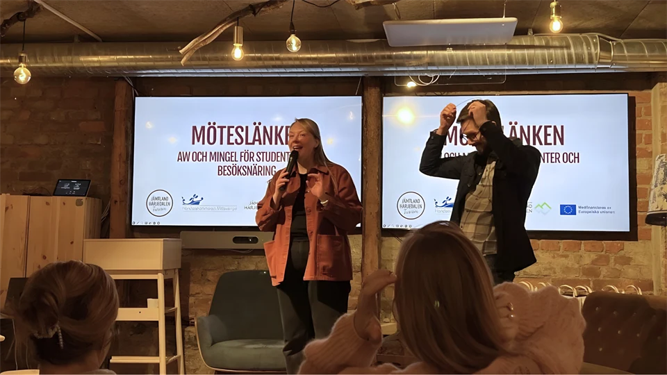 Kvinna och man på scenen under evenemanget Möteslänken 2025. Kvinnan pratar i en mikrofon som hon håller i.  