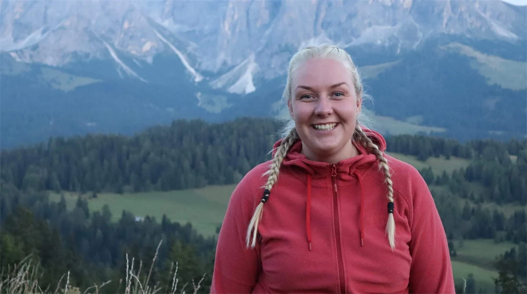 Kvinna i flätor och röd jacka i utomhusmiljö med höga berg i bakgrunden.  