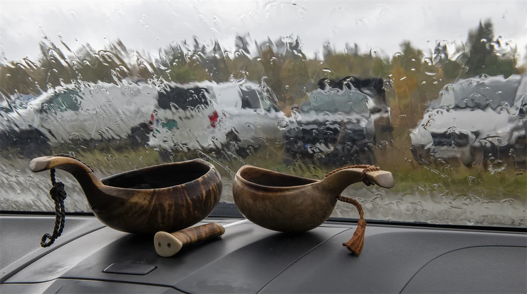 Två samiska kåsor och en samekniv i framrutan på en bil med intensivt regn utanför.