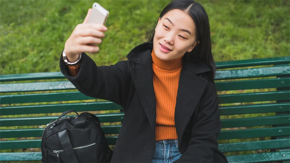 Kinesisk kvinna sitter på en bänk i en park och tar en selfie. Hon har på sig en orange tröja, svart jacka och jeans.