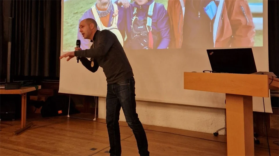 En man står på en scen och gestikulerar med en mikrofon i handen och en skärm med tre glada personer i utomhuskläder bakgrunden. 