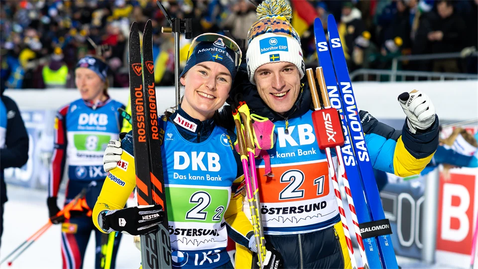 Två skidåkare bredvid varandra med tävlingskläder och skidor lutade mot axeln.