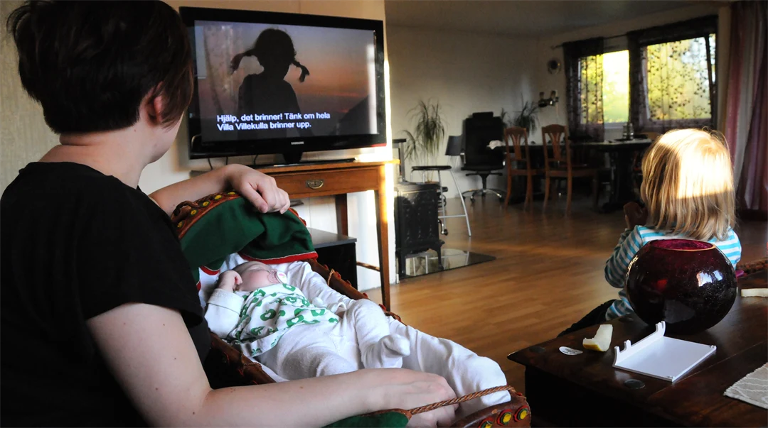 En kvinna och ett barn ser på Pippi Långstrump på TV. Kvinnan har en samisk vagga med en bebis i knäet.