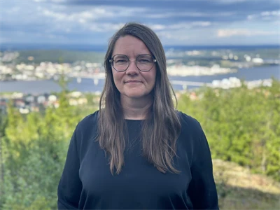 En kvinna i glasögn och långt, brunt hår klädd i en mörkblå långärmad tröja står framför ett naturlandskap med utsikt över träd och vatten.