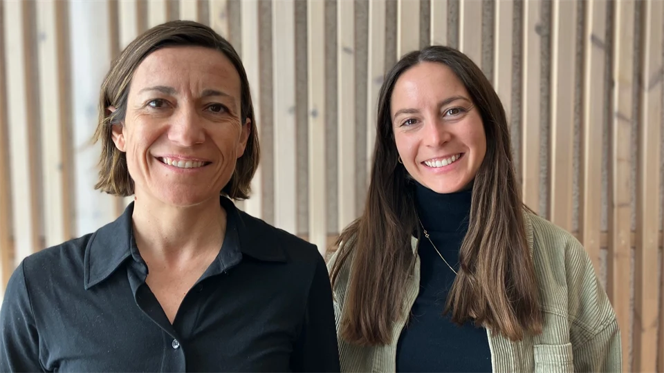 Two women stand in front of a wood-panelled wall and look into the camera, smiling