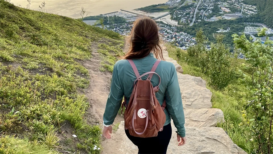 En kvinna med ryggsäck på ryggen går på en stenbelagd stig och ser ut över en norsk fjord.
