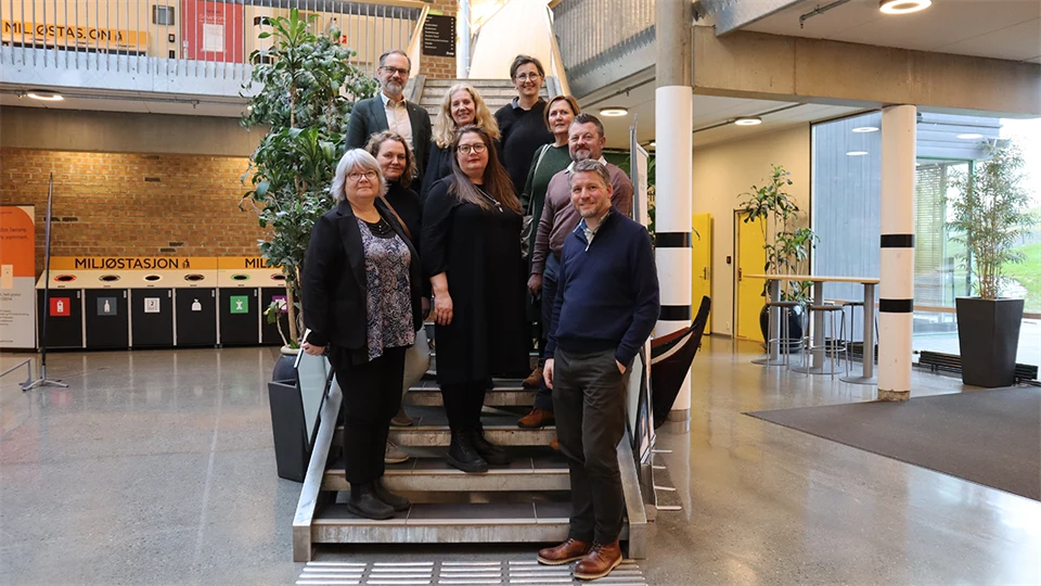 Nio personer står i inomhusmiljö i en trappa på de nedersta trappstegen. Runt omkring dem finns öppna ytor.