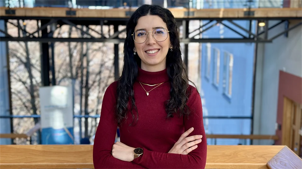 Smiling woman with dark curly hair, glasses, and a burgundy sweater standing with her arms crossed.