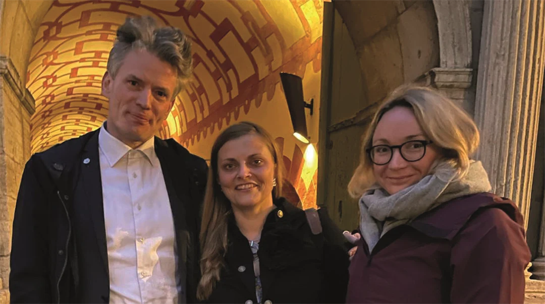 Three people, a man and two women, are standing in front of an illuminated stone archway.