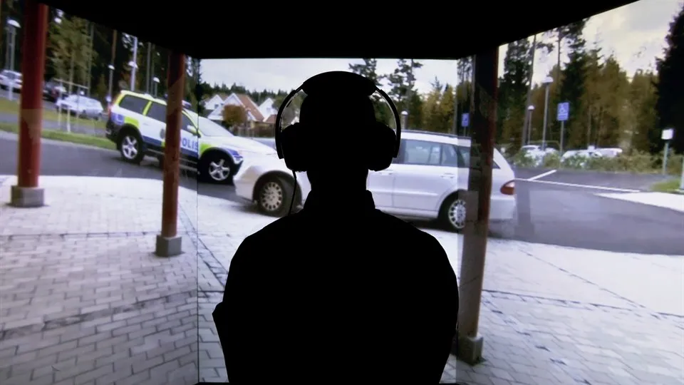 Siluetten av en person med hörlurar framför en stor skärm som visar en två bilar, varav en polisbil, vid en parkeringsplats.