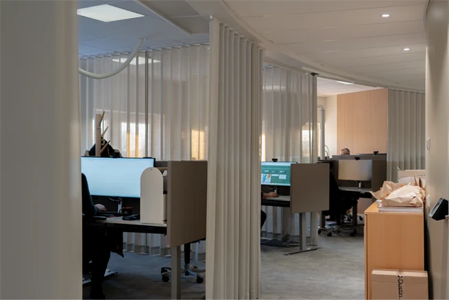 Upplyst kontorslandskap i ljust interiör uppdelat i olika sektioner av ljusgrå gardiner, tre personer sitter och arbetar vid egna skrivbord framför datorskärmar.