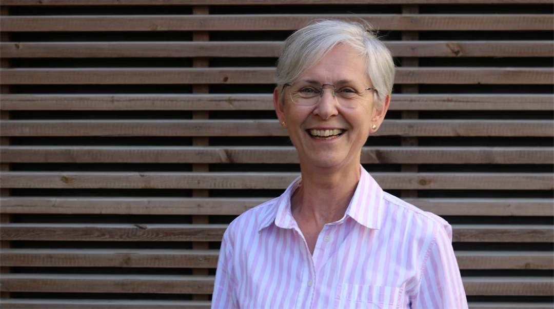 A woman with grey short hair, glasses and a pink and white striped shirt stands in front of a wall with horizontal wooden panelling.