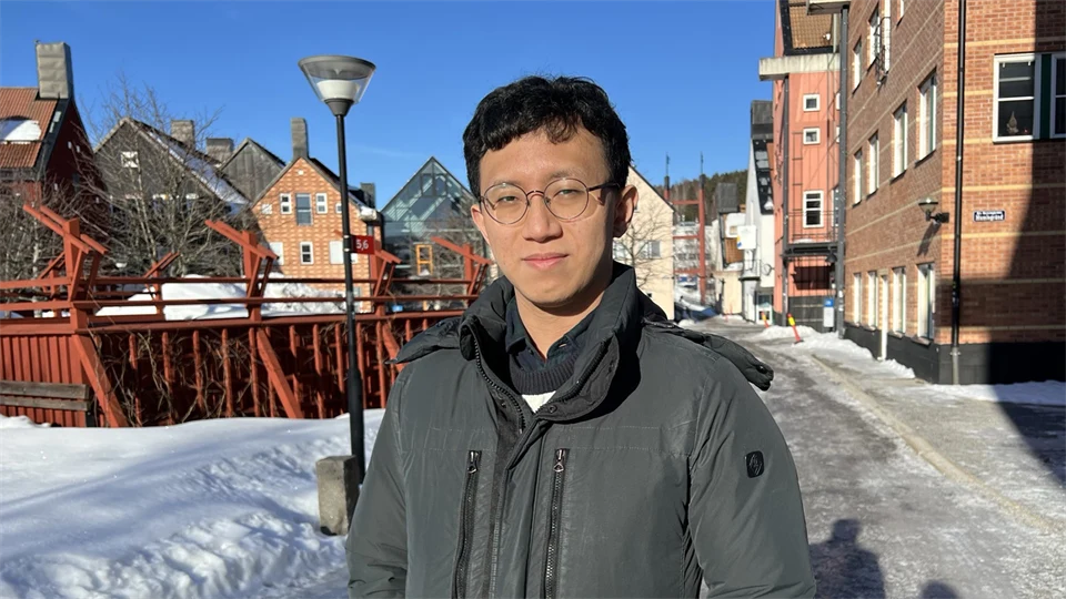 A man with dark hair and glasses stands outside in the sunshine. Campus buildings in snow and blue sky in the background.