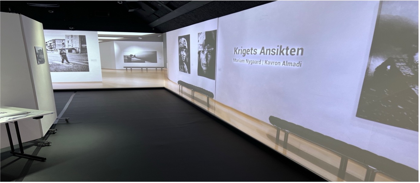 Utställningssal med projicerade fotografier och texten ”Krigets Ansikten” med två namn.