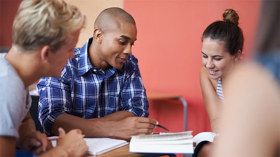 Grupp bestående av fyra studenter sitter och diskuterar runt en bok. Bakom dem syns en röd vägg och de ser glada ut.