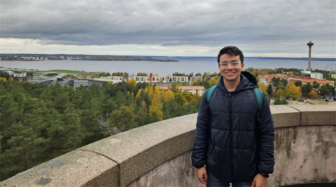 A young man wearing a dark blue jacket and carrying a rucksack is standing by a stone wall high up, looking out over trees, buildings and water. 