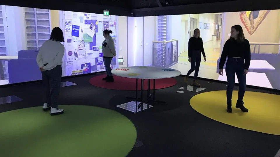 Four people are standing in a room. There are coloured circles on the floor. Some of the people are standing on the coloured circles. Pictures of a school environment are projected on the walls.