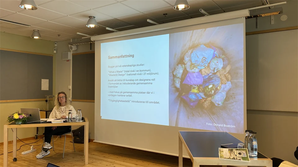 En kvinna i lockigt ljust hår sitter vid ett bord och håller en presentation som visas på en stor duk.