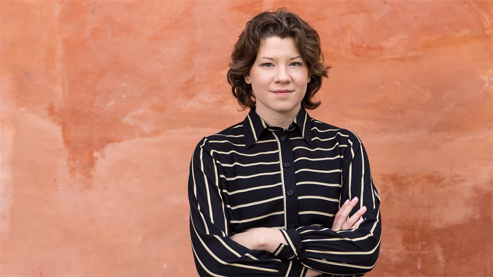 En kvinna med mörkt, kort, lockigt hår i en svart och vitrandig skjorta. Hon står mot en orange vägg.  