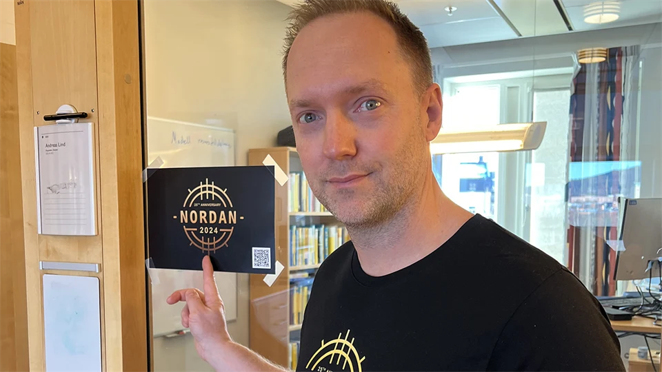 En man i svart t-shirt står och pekar på en dekal med texten Nordan2024.