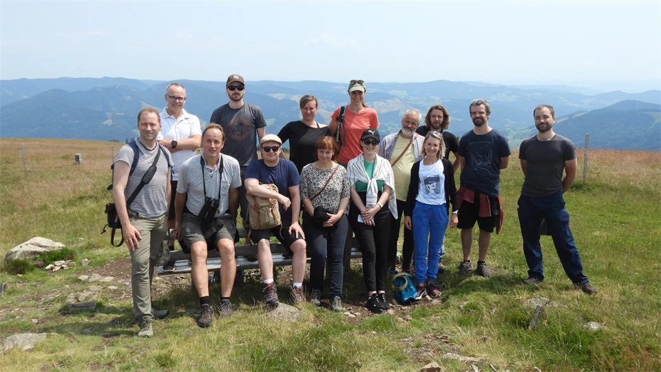  A group of people stand together in a meadow. A mountain landscape is visible in the background.