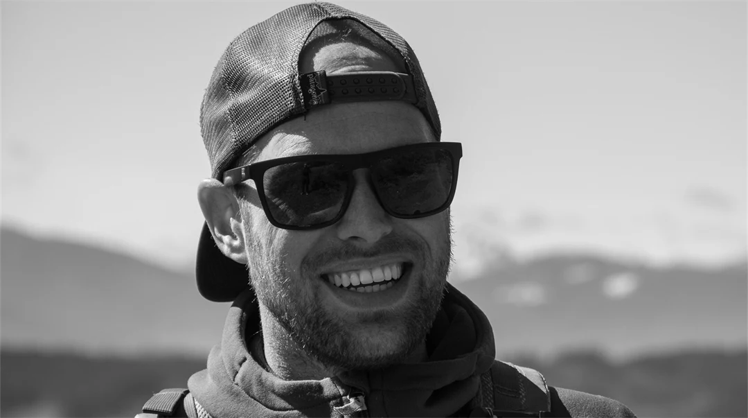 A black and white photo of young man standing in natural landscape with mountains in the background. He wears sunglasses and a cap back and front.