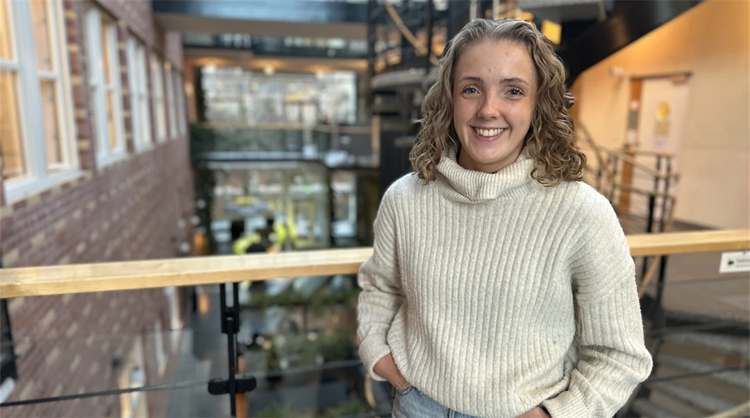 Ung kvinna med blont, lockigt, axellångt hår klädd i vit, stickad polotröja och ljusa jeans står i en ljus byggnad med många fönster, ett räcke och en trappa bakom sig. 