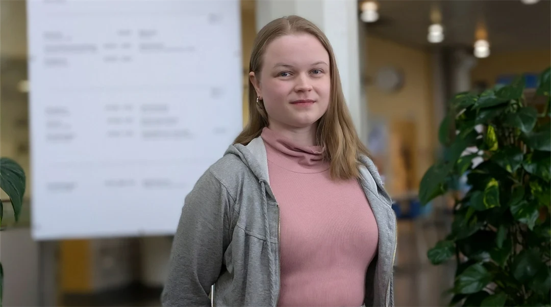 En kvinna med långt hår står framför en ljustavla. Bredvid finns en grön växt.