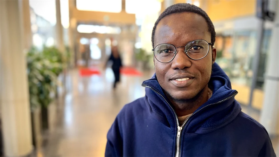 A man is standing in an hall. He is wearing a blue hoodie.
