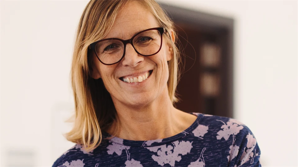 En person med axellångt blont hår står inomhus i en ljus miljö. Personen bär en mörkblå tröja med ett mönster av ljusa blommor.