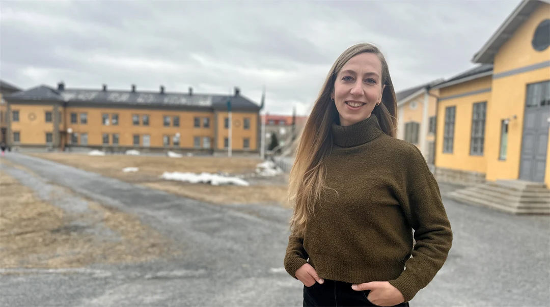 Kvinna i brun polotröja och svarta byxor står utomhus framför gula stora byggnader