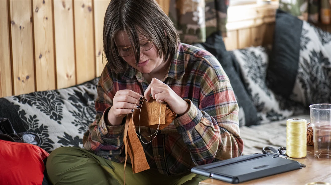 En ung person sitter i en soffa och syr i renskinn