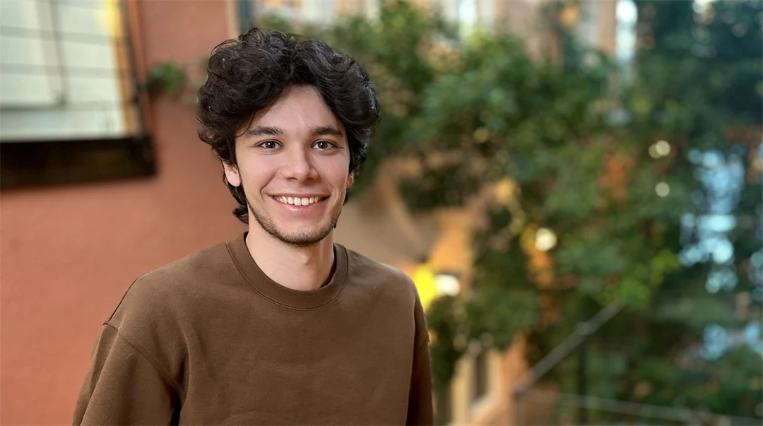 A young man with black curly hair and a brown sweater. In the background, green vegetation and stone walls in red and beige can be seen. 