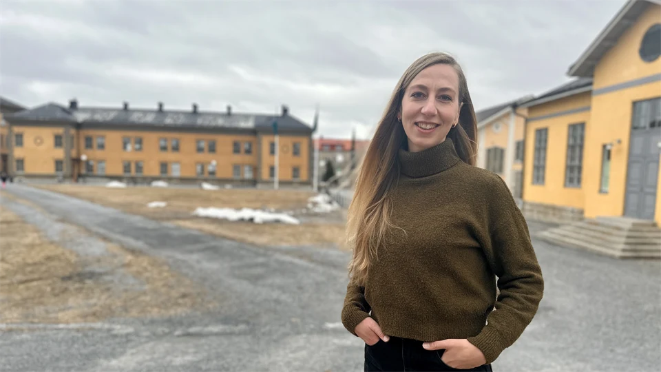 Woman in brown polo shirt and black trousers standing outdoors in front of large yellow buildings
