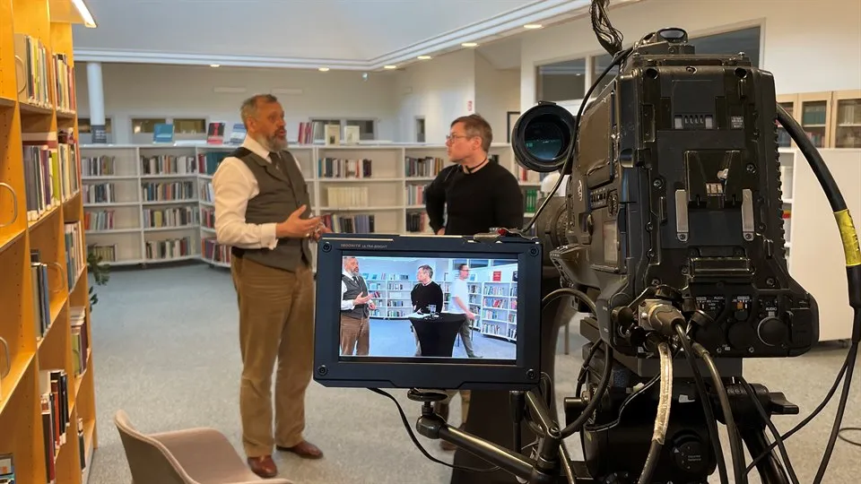 Två män står i ett bibliotek och pratar med varandra. I förgrunden syns en filmkamera. Männen syns även på kamerans skärm.